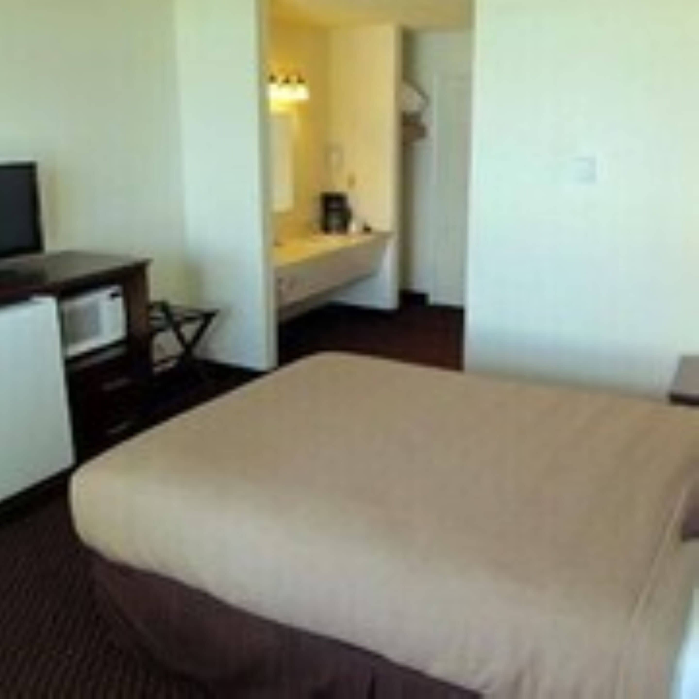 Une chambre d’hôtel avec un lit, un bureau équipé d’un ordinateur, un petit réfrigérateur et une salle de bain visible à travers une porte ouverte.