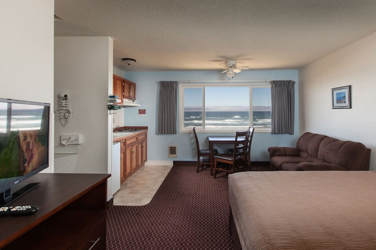 Quarto de hotel com cama, televisão, cozinha compacta, mesa de jantar com cadeiras e vista para o oceano.