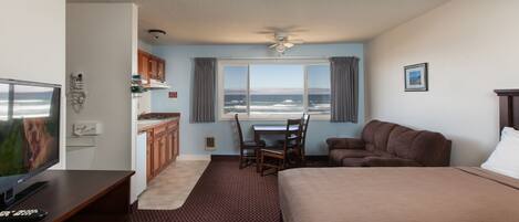 A hotel room with a bed, a TV, a kitchenette, a dining table with chairs, and a view of the ocean.