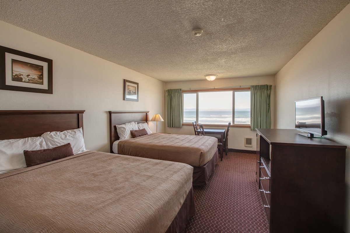 Quarto de hotel com duas camas, uma escrivaninha, televisão e janela com vista para o oceano.
