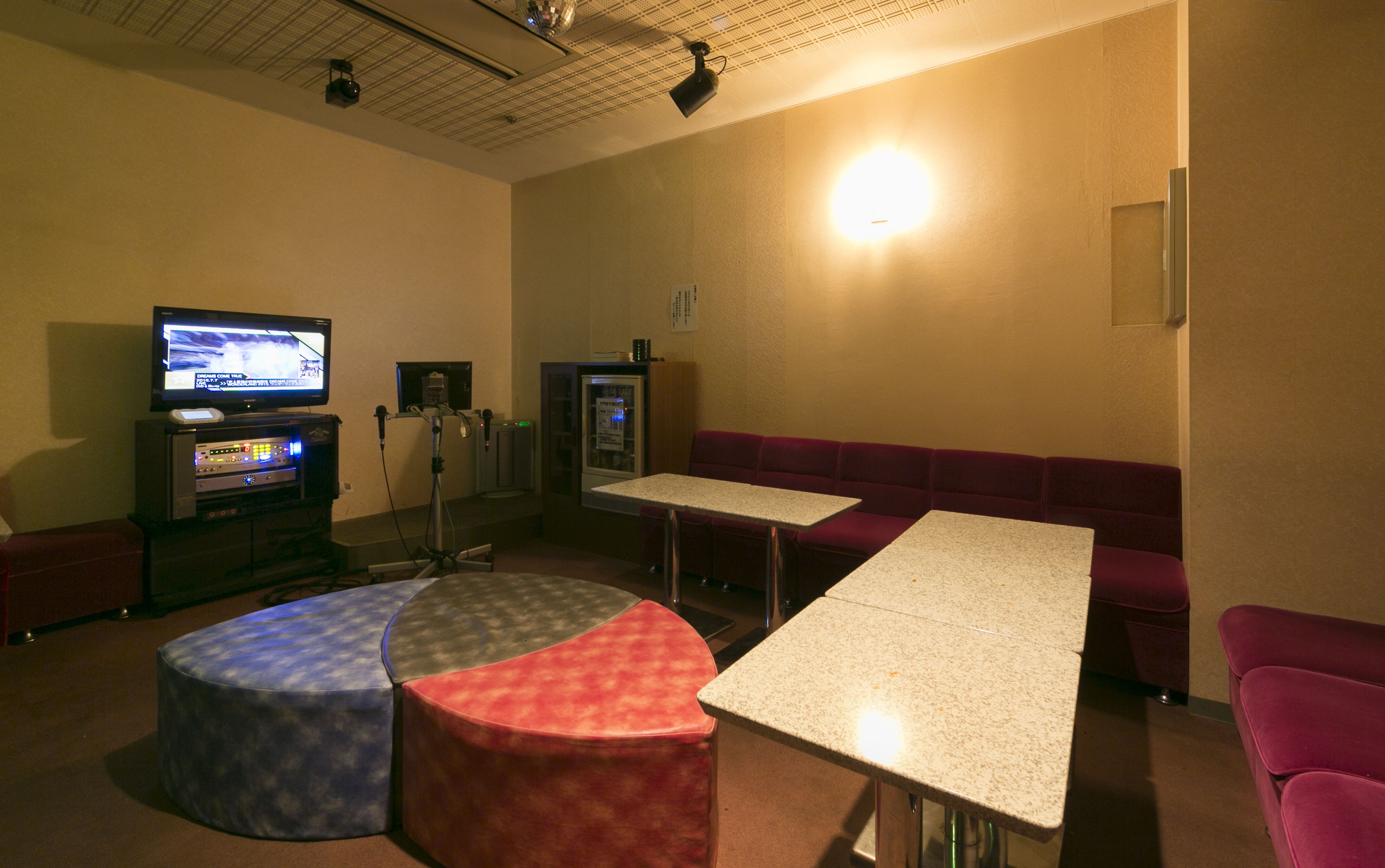 Quarto com televisão, pequena geladeira, mesa, sofá e um banquinho redondo.