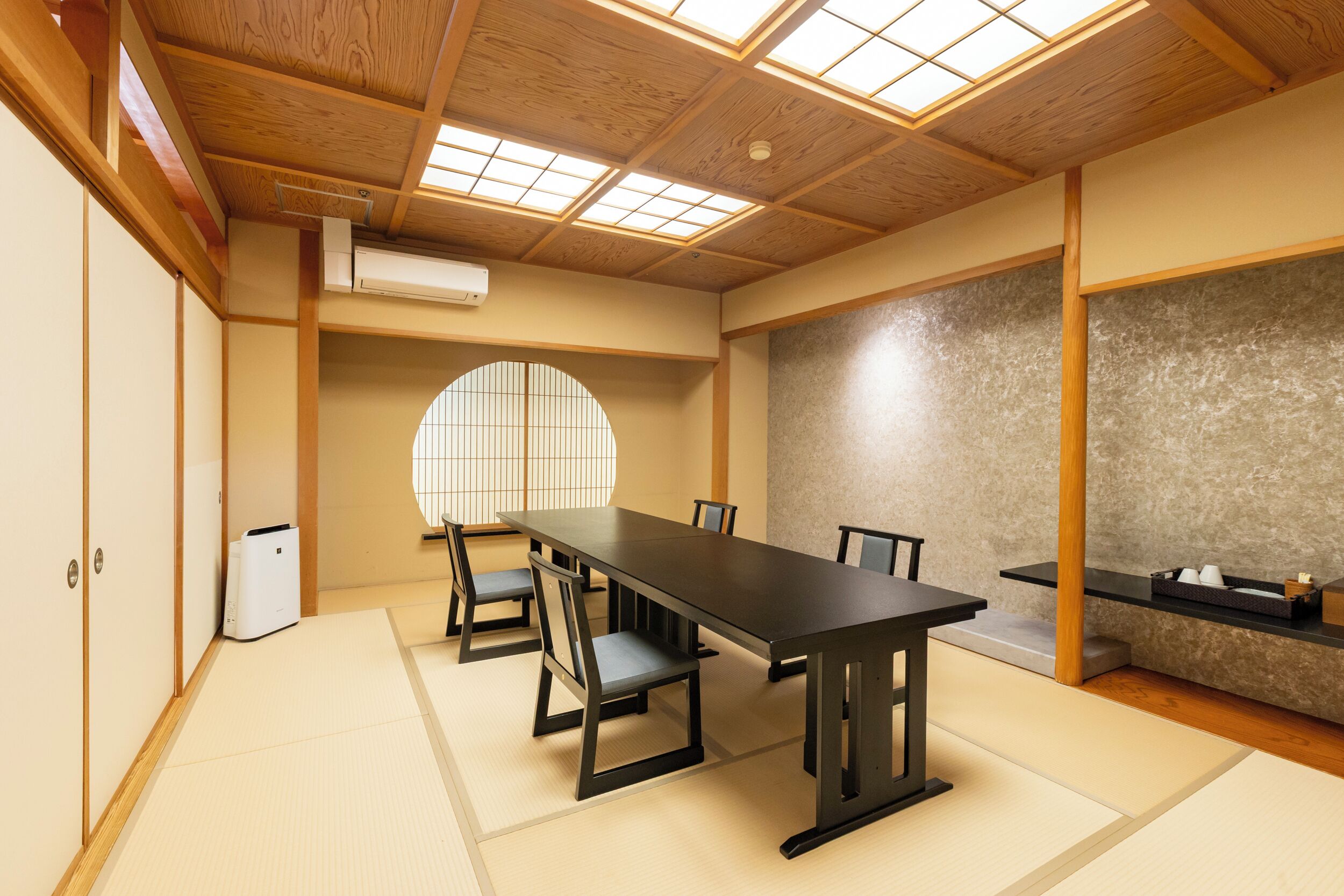 A traditional Japanese room with tatami flooring, a low table with chairs, a large window, and a stone wall.