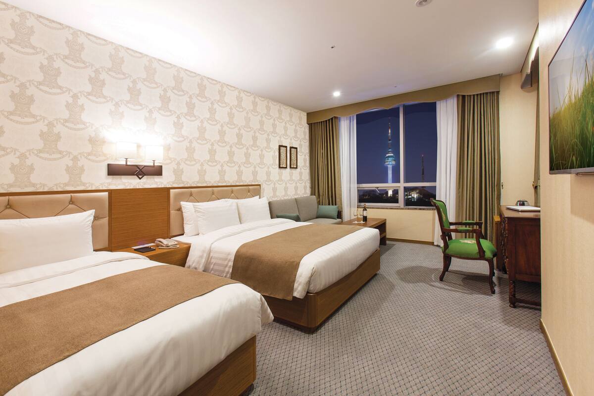 A hotel room with two beds, a desk, a chair, a window with a cityscape view, and a painting on the wall.