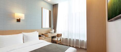 Habitación de hotel con una cama grande, un escritorio, una silla, una lámpara de escritorio y una ventana con cortinas translúcidas.
