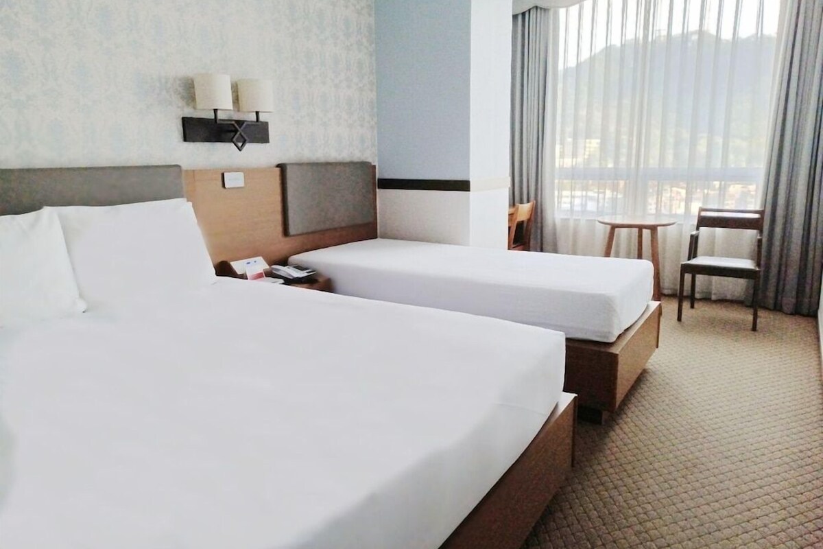 Habitación de hotel con dos camas, una mesita de noche, una lámpara, una silla y una ventana con cortinas.