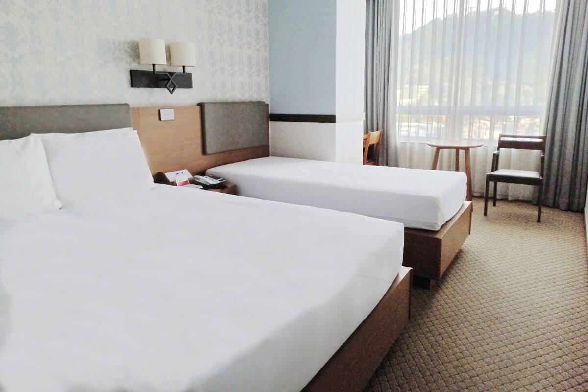 A hotel room with two beds, a nightstand, a lamp, a chair, and a view of the outdoors through sheer curtains.