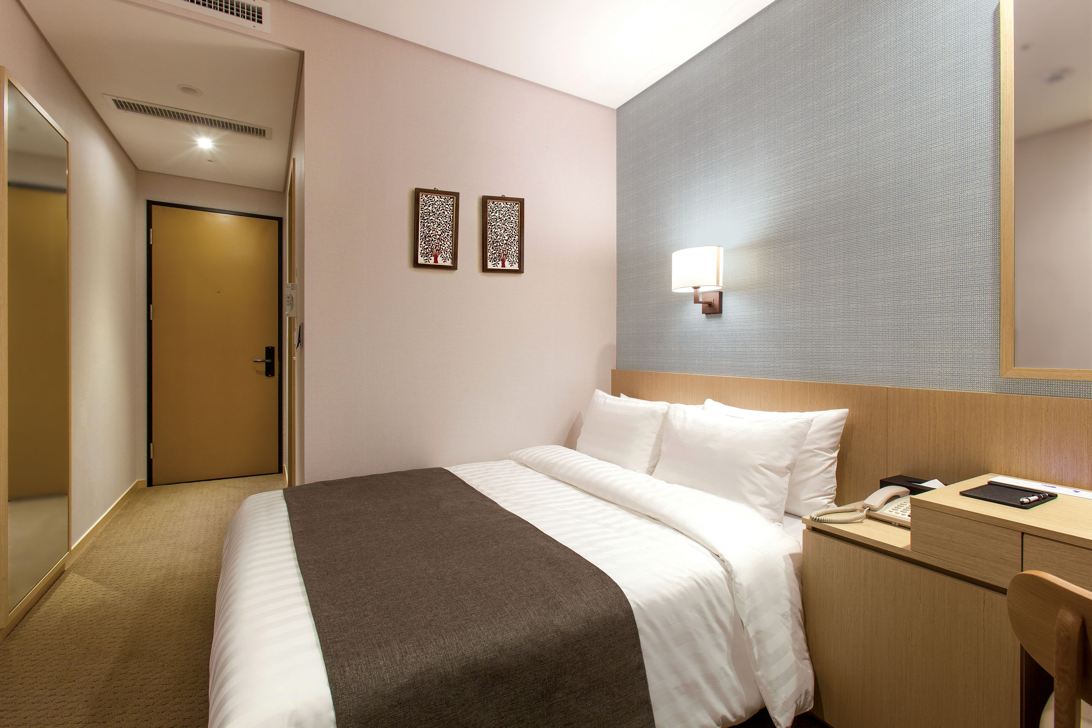 Ein Hotelzimmer mit einem Bett, einem Schreibtisch mit Telefon und Tablet, einem Spiegel und zwei Wandleuchten.