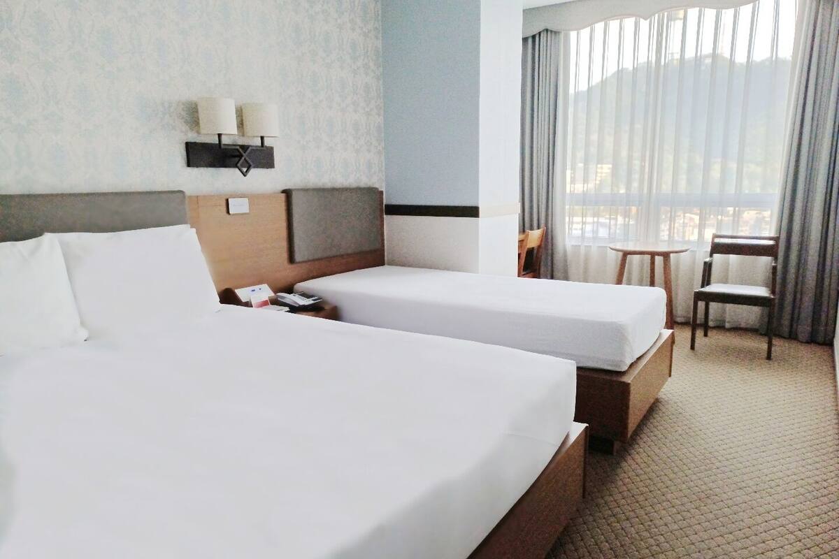 A hotel room with two beds, a nightstand, a lamp, a chair, and a view of the outdoors through sheer curtains.