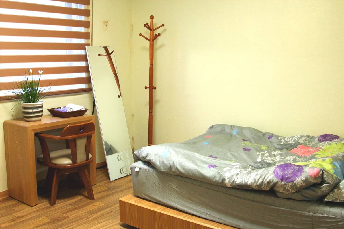 A bedroom with a bed, a desk, a chair, a coat rack, and a window with blinds.