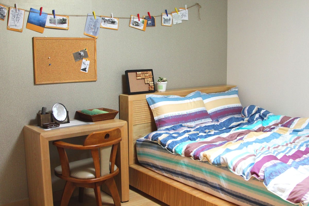A bedroom with a bed, a desk, and a chair. There is a corkboard on the wall with photos and notes pinned to it.