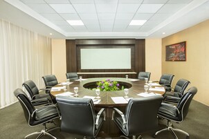 Uma sala de conferências com uma mesa redonda, cadeiras de couro preto, uma tela de projetor e um arranjo floral.