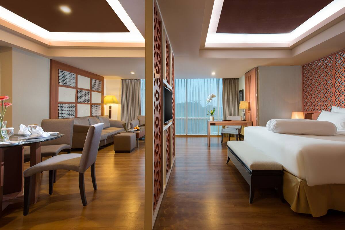 A hotel room with a sofa, a dining table with chairs, a TV, a bed, and a view of the city.