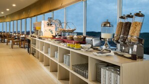 Een buffetopstelling met diverse gerechten en bestek, een koffiemachine en uitzicht op de zee door grote ramen.