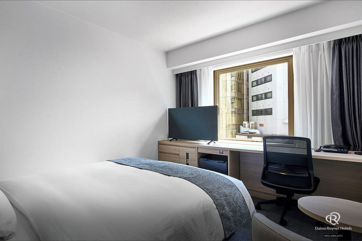 Quarto de hotel com cama, escrivaninha com computador, cadeira, televisão e janela com cortinas.