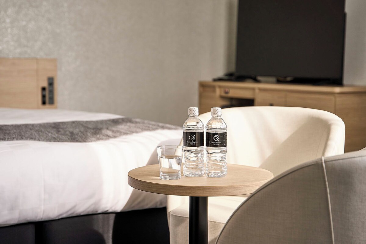 A hotel room with a bed, a round wooden table with two water bottles and a glass, a chair, and a TV on a wooden cabinet.