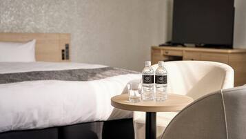 A hotel room with a bed, a round wooden table with two water bottles and a glass, a chair, and a TV on a wooden cabinet.