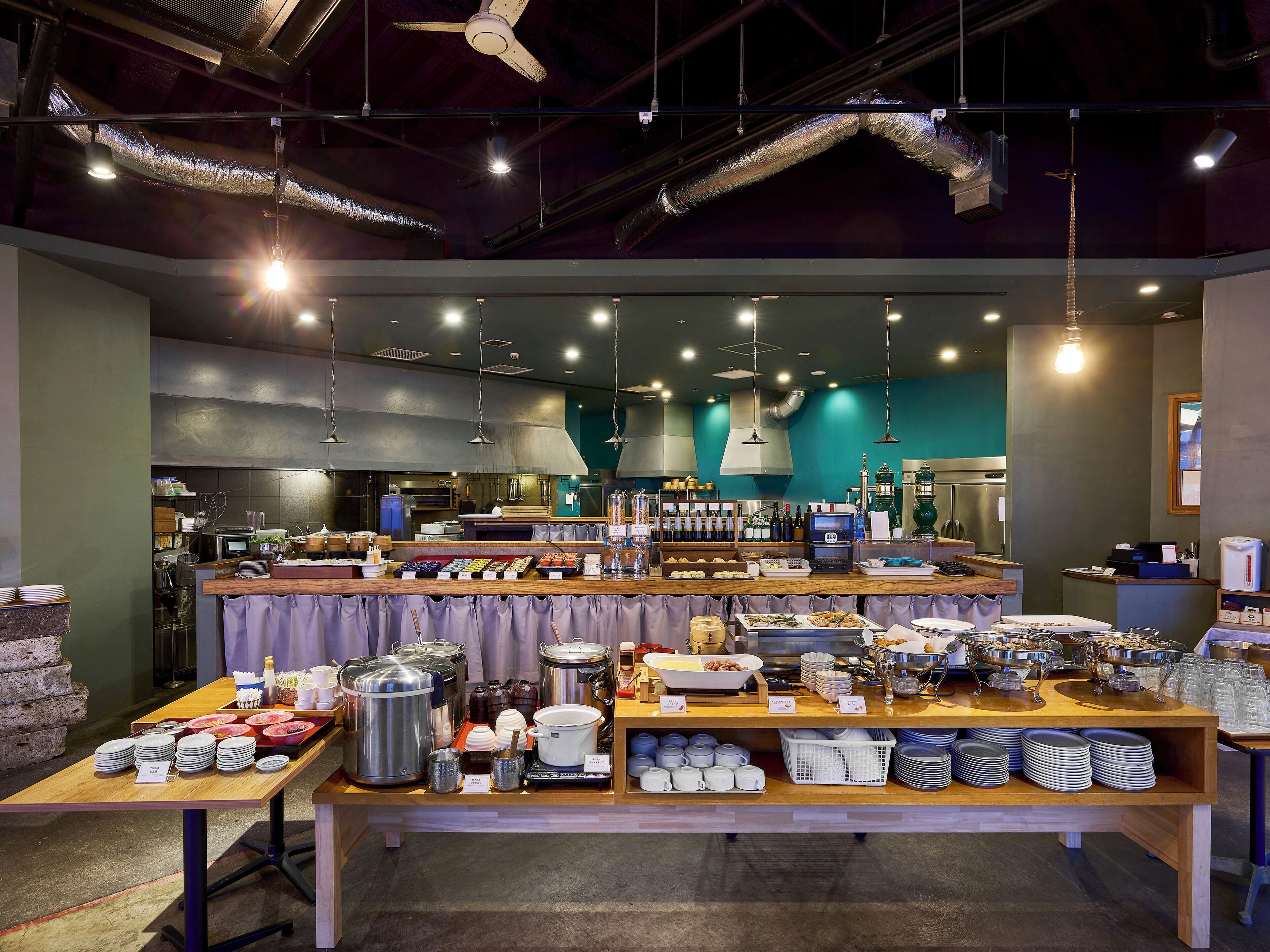 A modern cafe with a central serving counter, multiple seating areas, and a visible kitchen in the background.