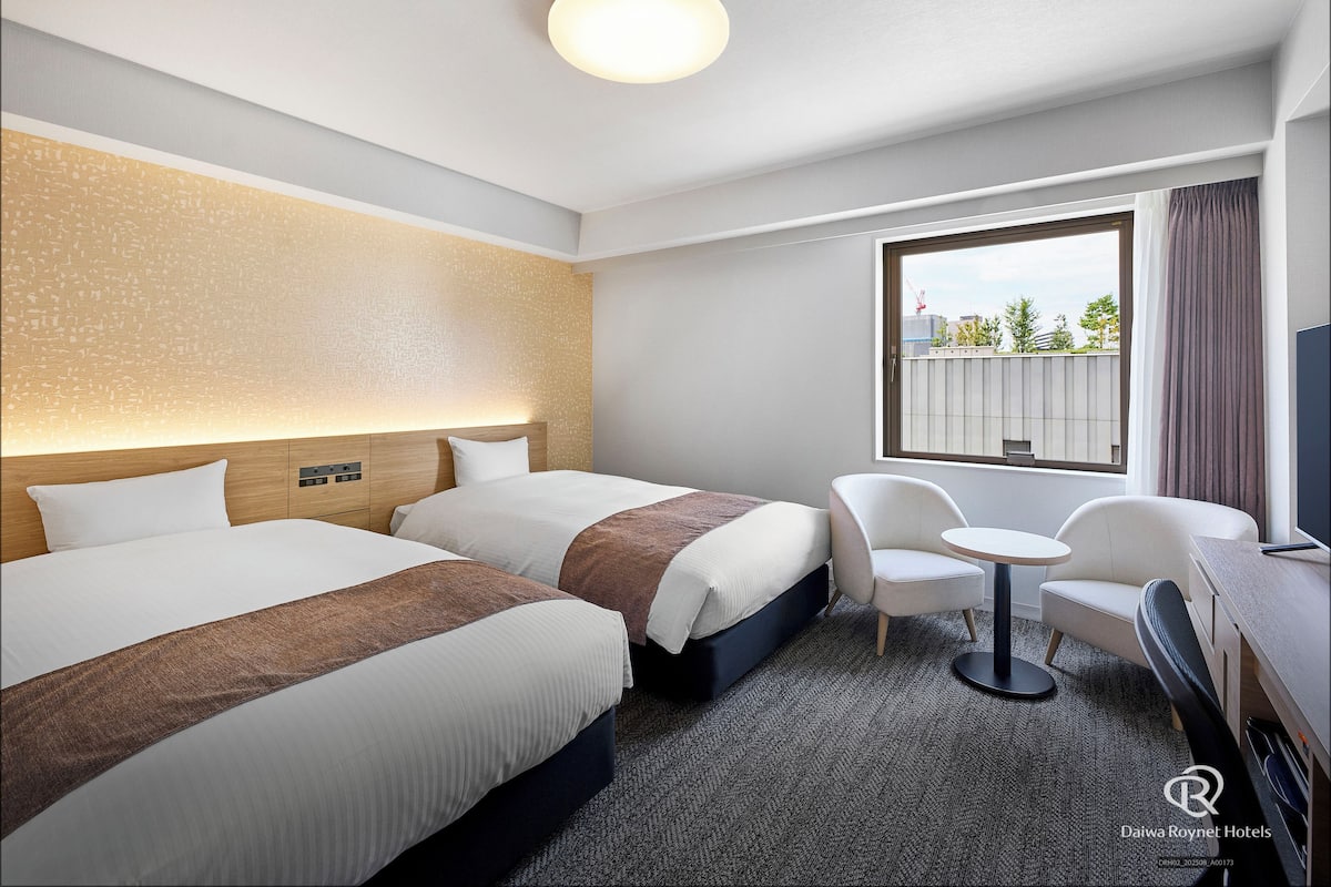 Quarto de hotel com duas camas, uma mesa redonda pequena, uma cadeira e uma televisão.