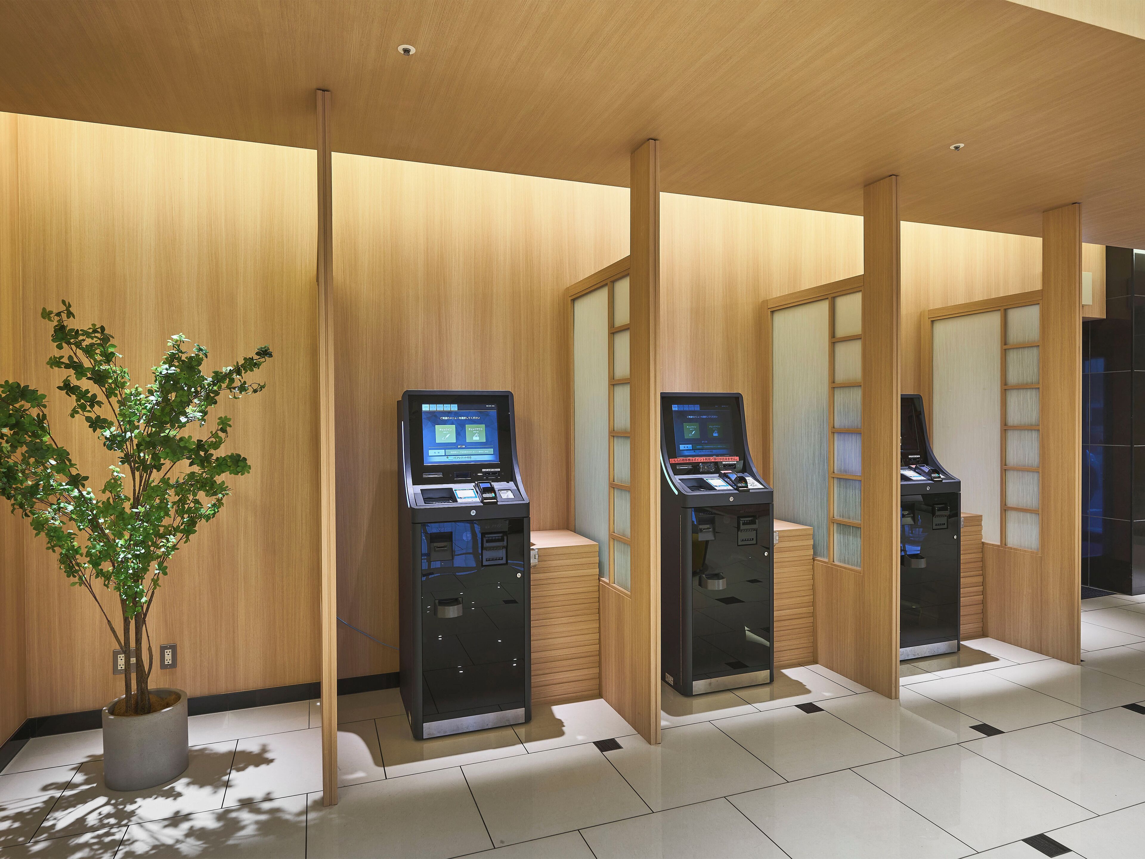 Un moderno vestíbulo de hotel con paneles de madera, una planta en maceta y quioscos de autoservicio.