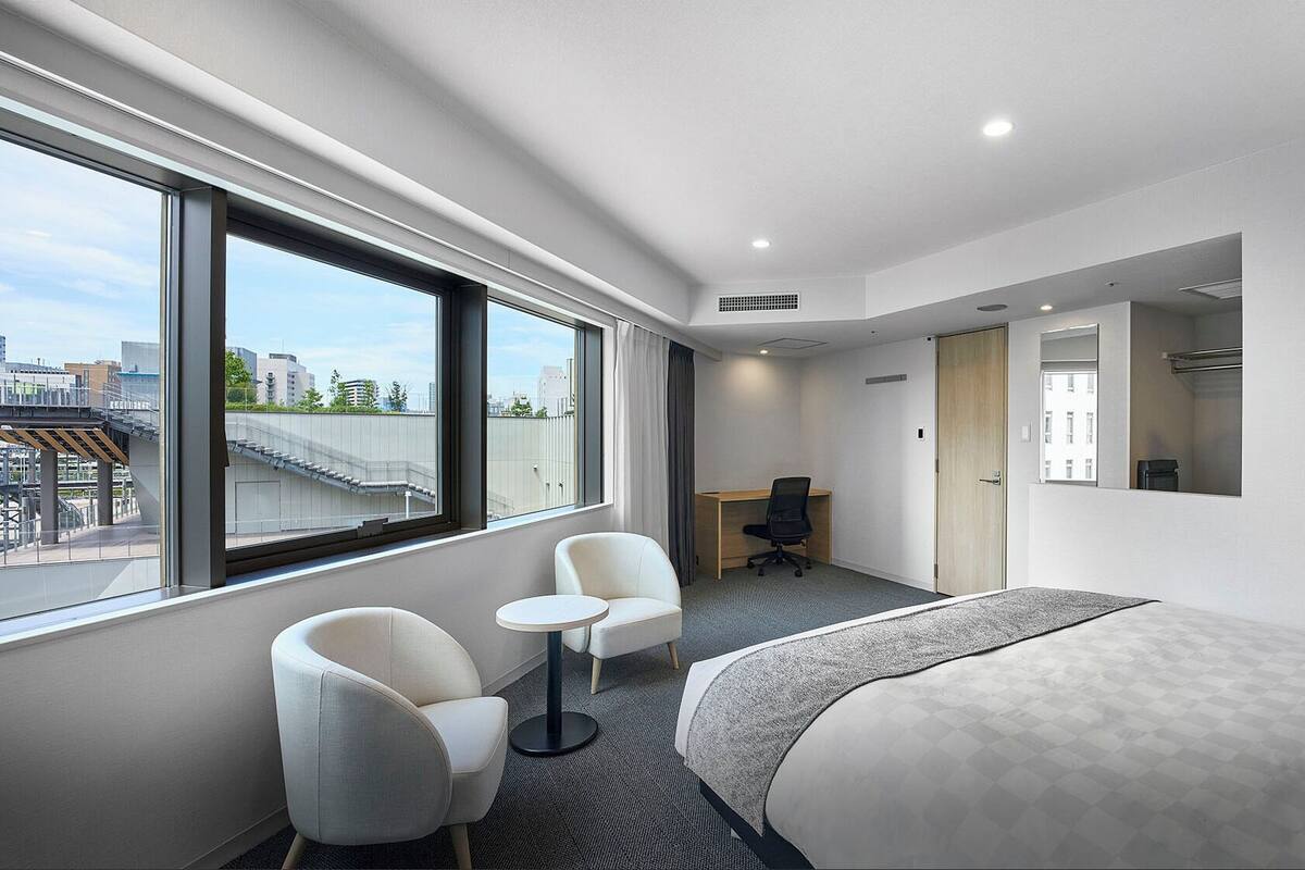 Quarto de hotel moderno com uma janela grande, duas cadeiras, uma escrivaninha e uma cama.