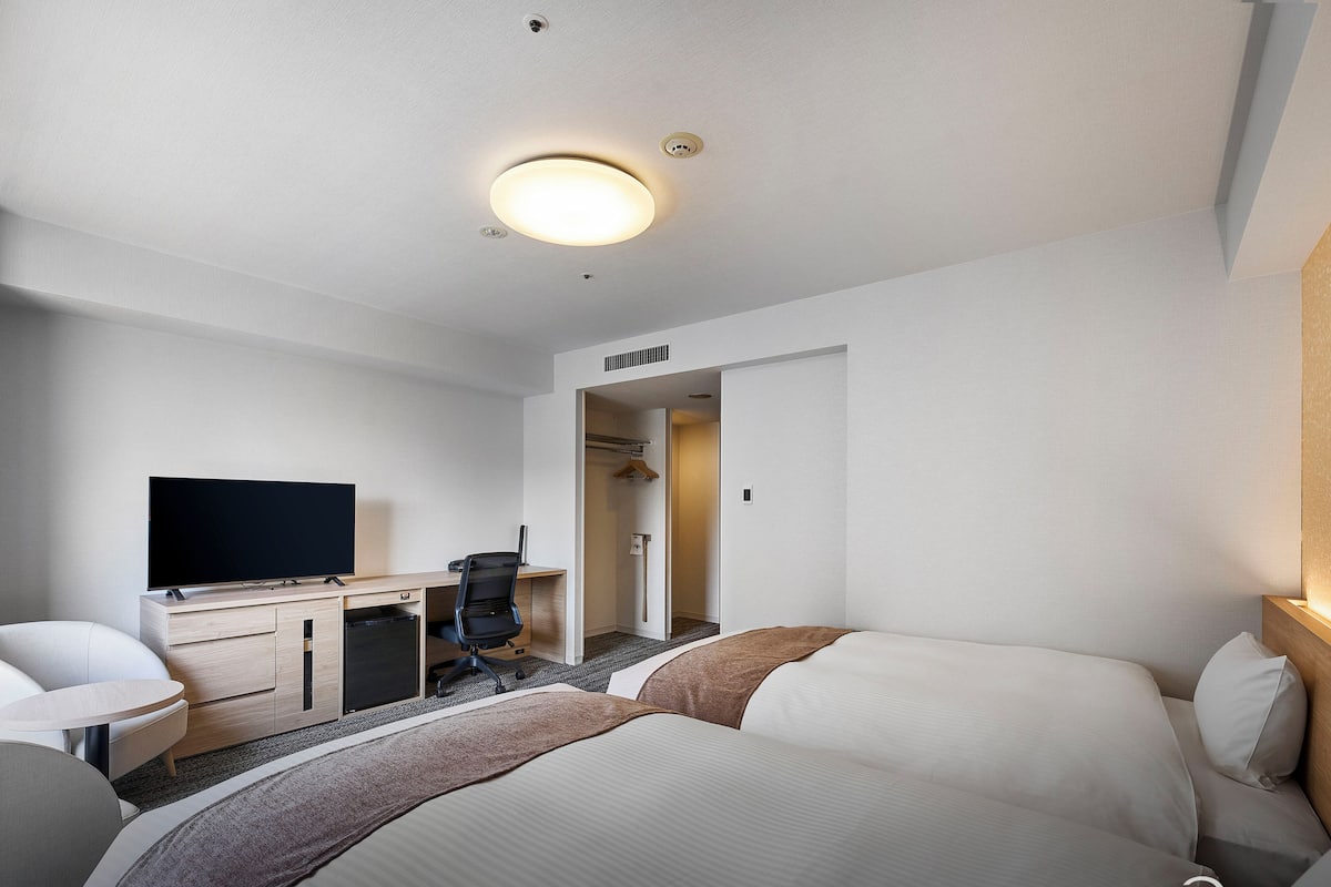 Quarto de hotel com cama, escrivaninha com televisão, cadeira e closet espaçoso.