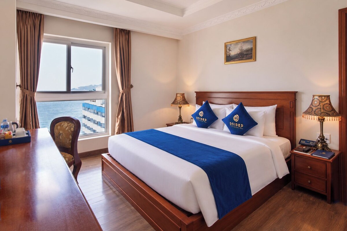 Een hotelkamer met een groot bed, twee nachtkastjes, een bureau en uitzicht op zee.