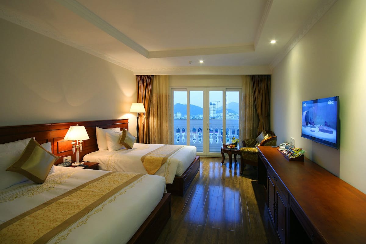 Ein Hotelzimmer mit zwei Betten, einem Fernseher, einem Schreibtisch und Blick auf die Skyline durch große Fenster.