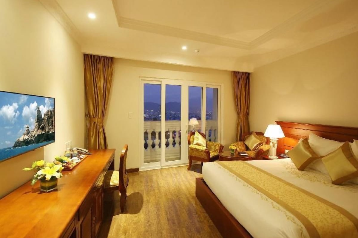 A hotel room with a bed, a desk, a chair, a TV, and a balcony with curtains.
