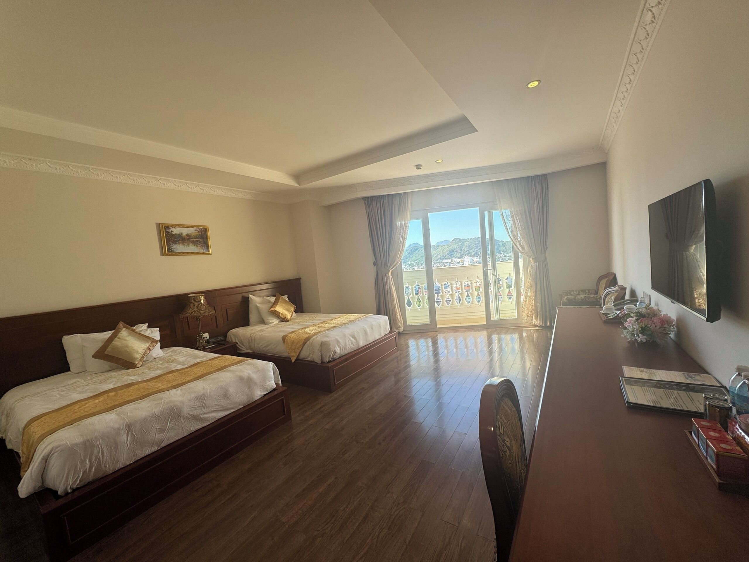 A hotel room with two beds, a large desk, a TV, and a balcony with a view.