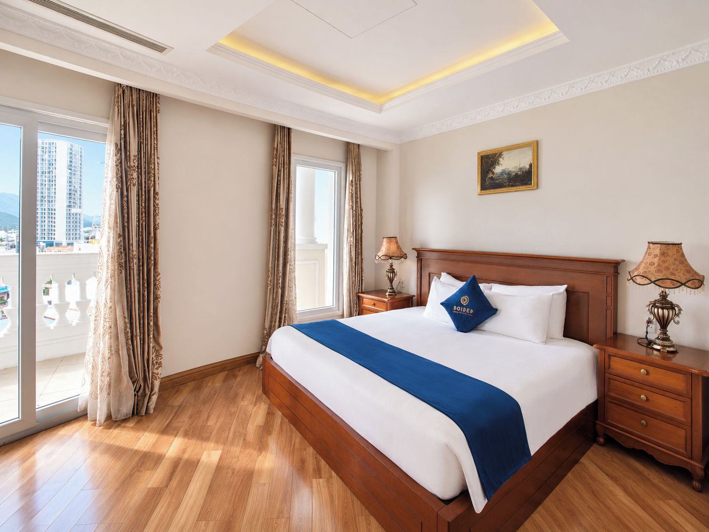 A hotel room with a large bed, wooden headboard, two bedside tables with lamps, a painting on the wall, and a balcony with curtains.