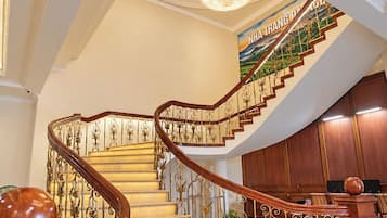 Un magnifique escalier orné de balustrades travaillées et surmonté d’un lustre, menant à une pièce aux murs lambrissés et où se trouve un piano.