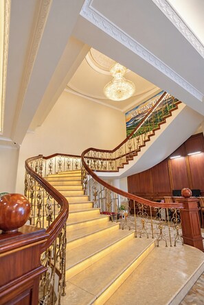 Un magnifique escalier orné de balustrades travaillées et surmonté d’un lustre, menant à une pièce aux murs lambrissés et où se trouve un piano.