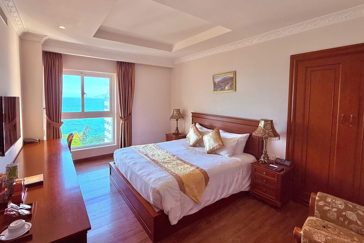 Ein Hotelzimmer mit einem großen Bett, Holzmöbeln, einem Schreibtisch mit Annehmlichkeiten, Meerblick und einem Gemälde an der Wand.