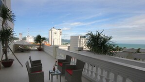 Een balkon met rieten stoelen en een kleintje tafeltje, met uitzicht op een stadsgezicht met gebouwen en een waterpartij.