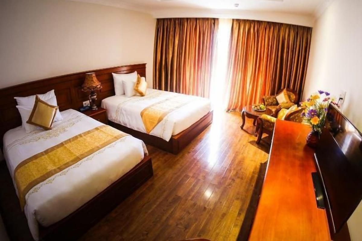 A hotel room with two beds, a wooden floor, a desk with a flat-screen TV, and a view of the outdoors through curtains.