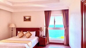 Une chambre d’hôtel avec un grand lit, une table de chevet, une lampe, un tableau au mur et une vue sur la mer par la fenêtre.