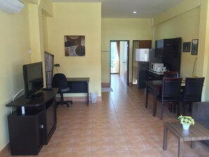 A modern living room with a flat-screen TV, a black office chair, a dining table with chairs, and a kitchen area with a refrigerator and microwave. - Nin Apartments Karon Beach (Karon)
