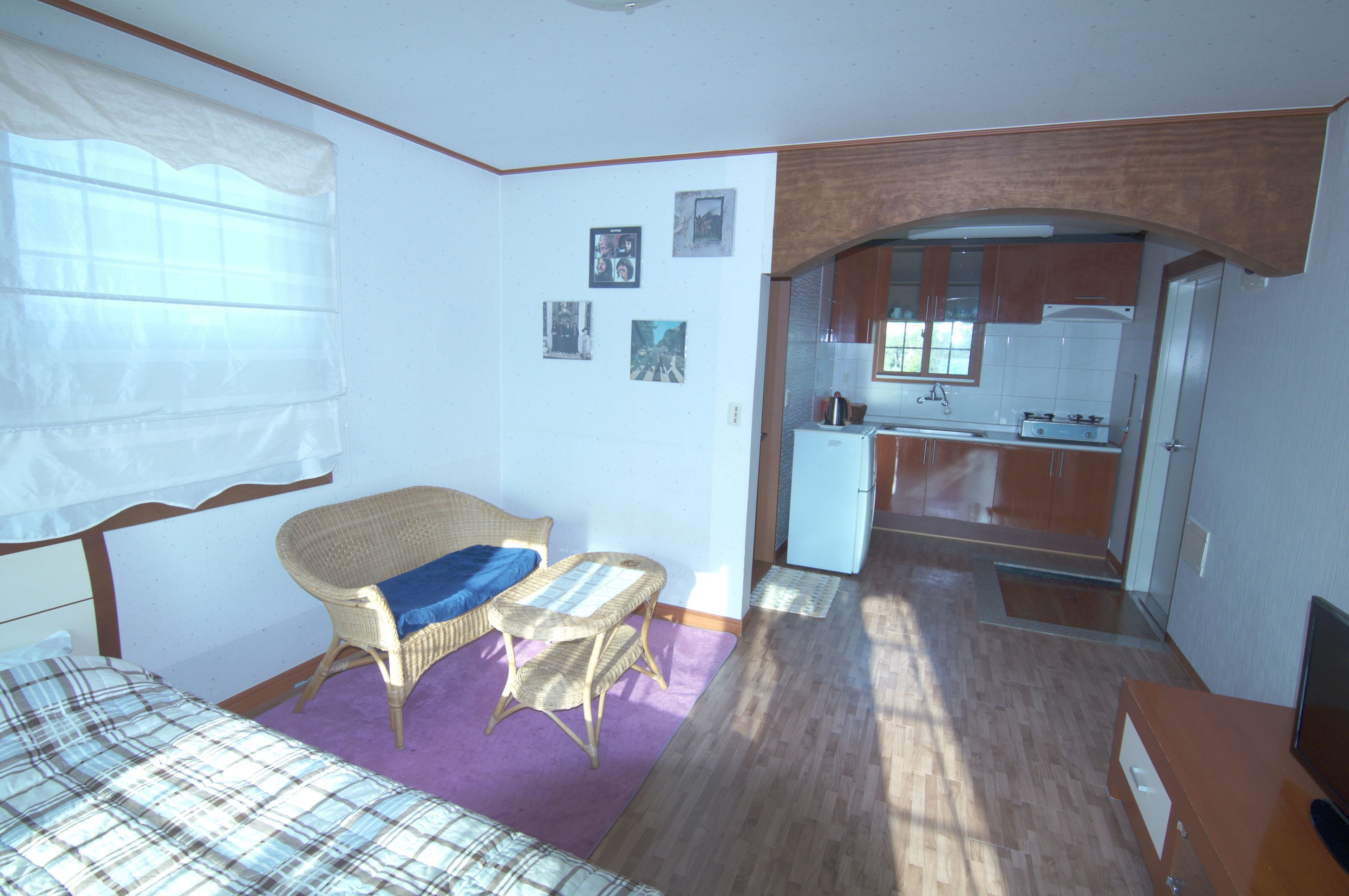 A compact room with a bed, a wicker chair, a small table, a kitchenette, and a window with sheer curtains.