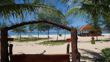 Un banco de madera bajo una palmera en una playa arenosa, con una cabaña de techo de paja al fondo.