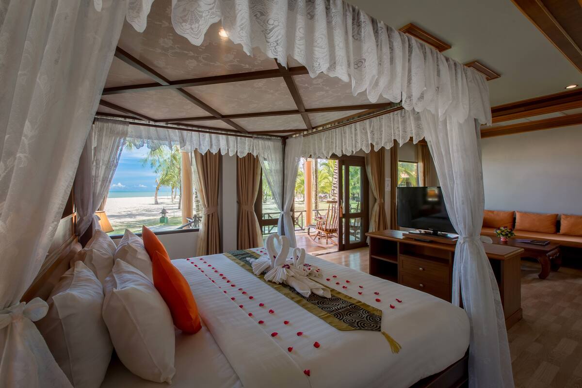 A bedroom with a bed, curtains, a TV, and a view of the beach.
