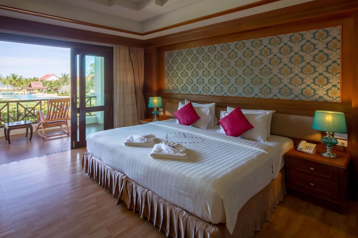 A hotel room with a large bed, two bedside tables with lamps, a view of the pool, and a balcony with a chair.