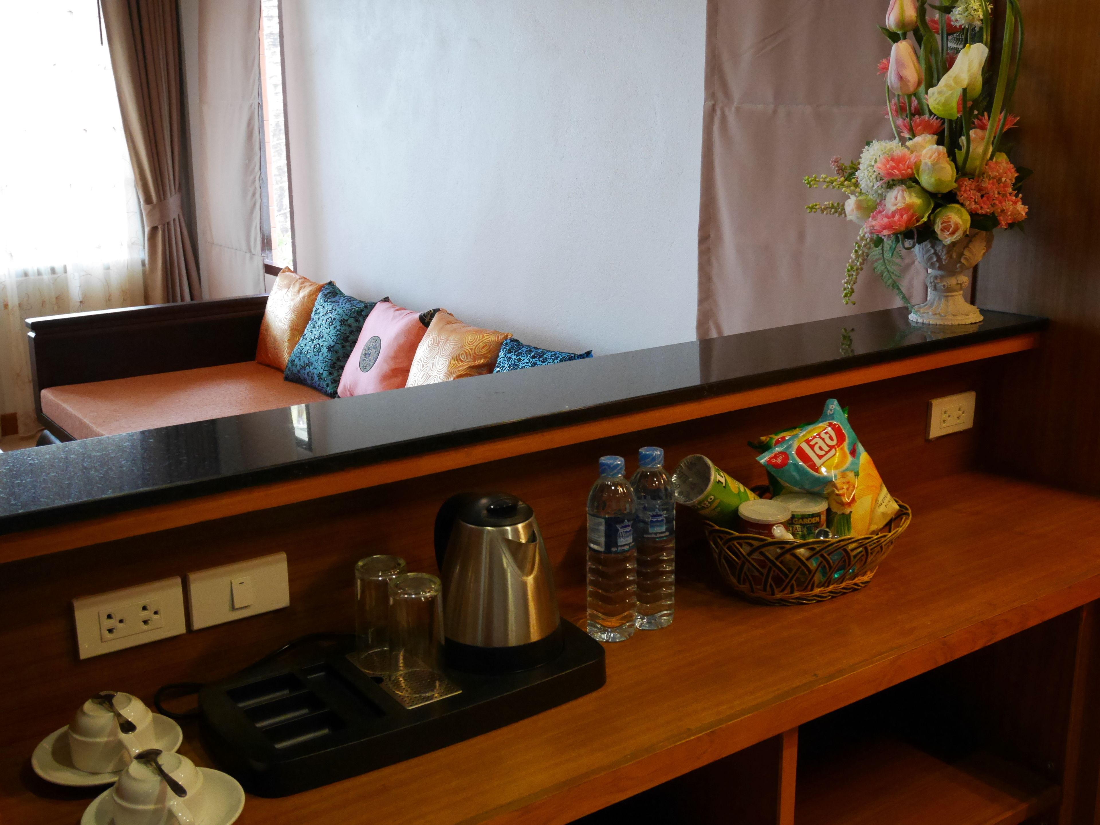 A hotel room with a wooden bench, pillows, a vase of flowers, a coffee maker, water bottles, a basket with snacks, and a tea set.