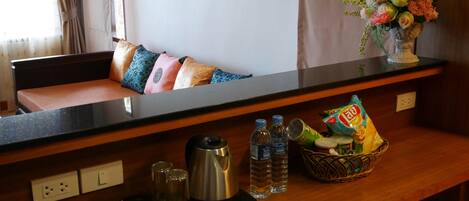 A hotel room with a wooden bench, pillows, a vase of flowers, a coffee maker, water bottles, a basket with snacks, and a tea set.
