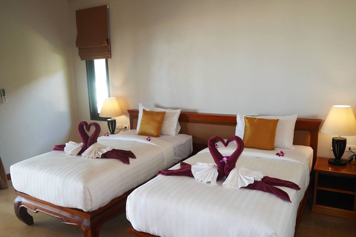 Two neatly made beds with white linens and decorative swan-shaped towels, a wooden headboard, bedside lamps, and a window with a blind.