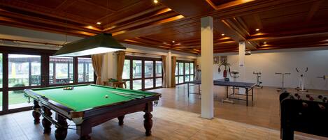 A spacious indoor area with a pool table, exercise equipment, and large windows offering views of the outdoors.