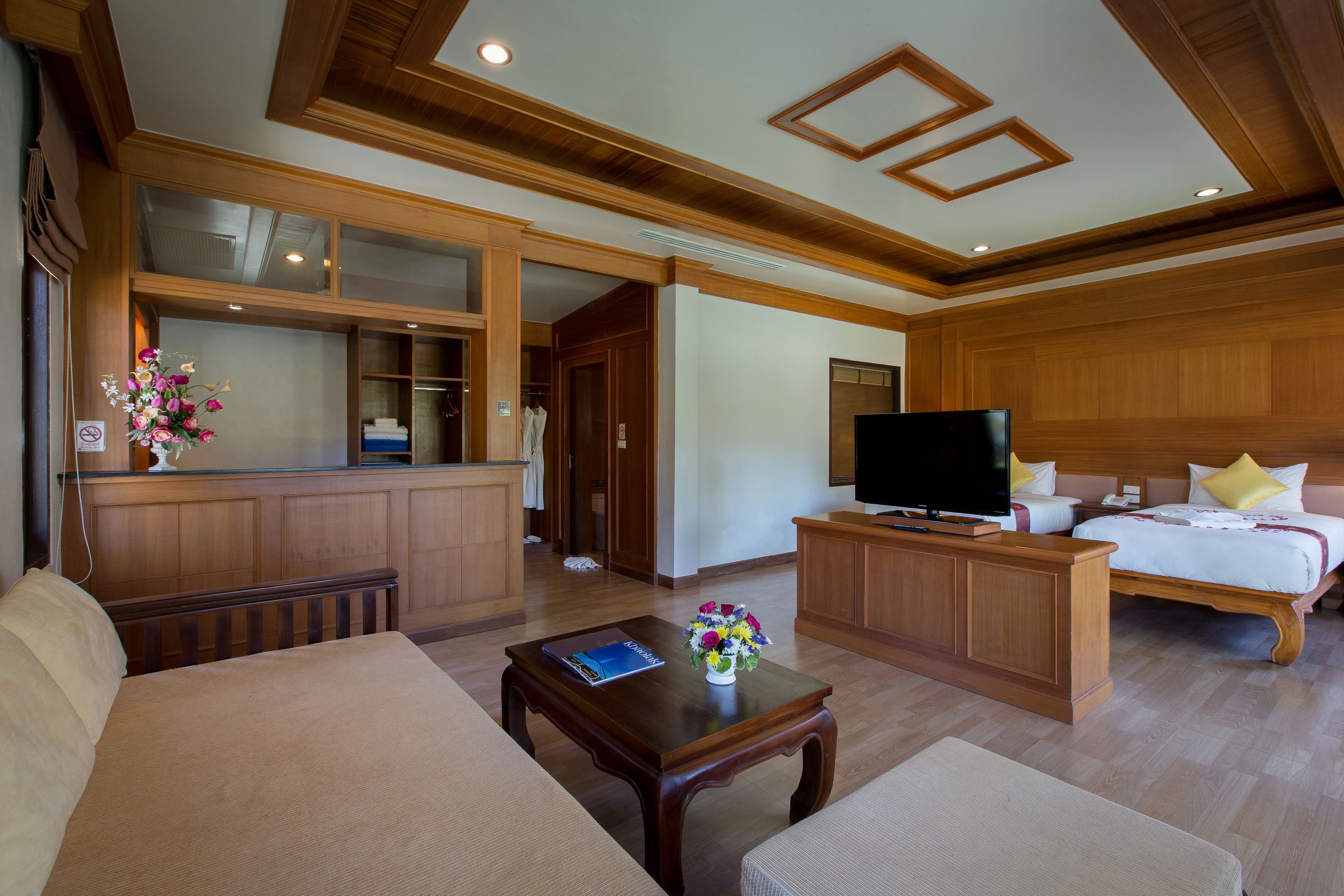 Ein Hotelzimmer mit Holzboden, einem Fernseher auf einem Holzständer, einem Bett mit weißer Bettwäsche und gelben Kissen, einem Holztisch und einer Blumenvase.