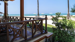 A beach view from a covered area with a wooden railing, palm trees, and beach umbrellas.