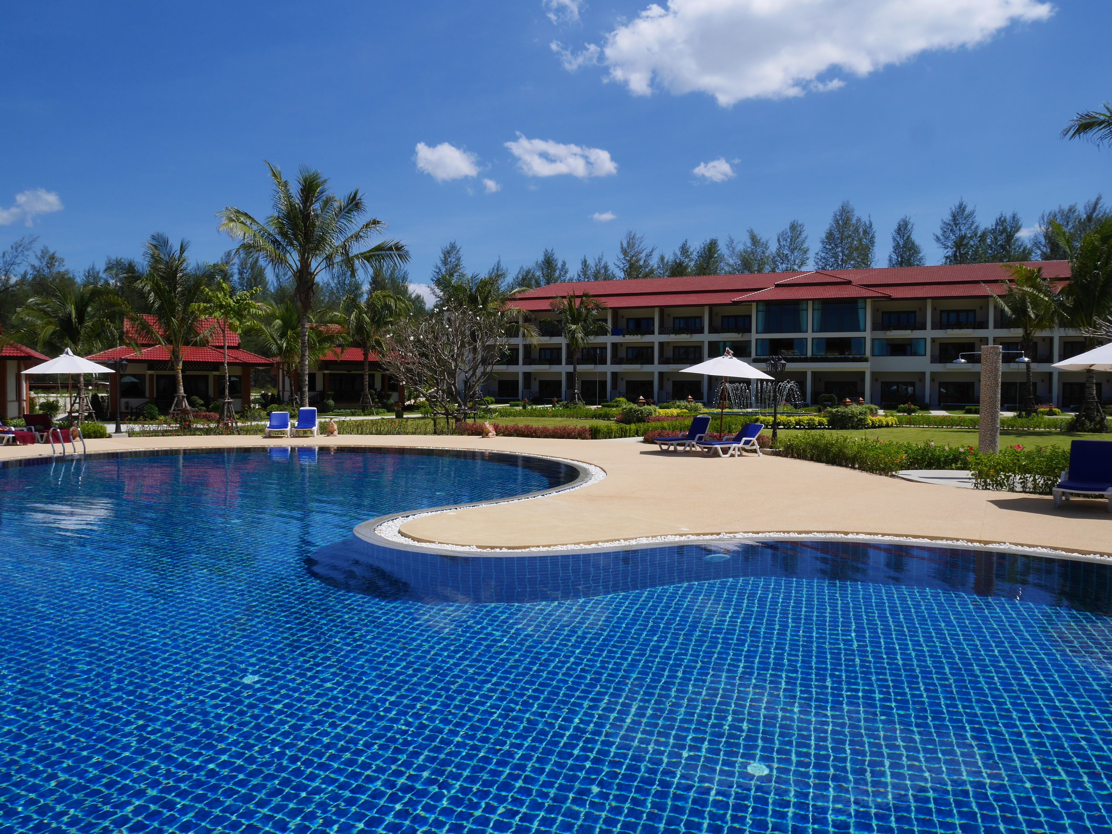 A swimming pool with lounge chairs and umbrellas, surrounded by a landscaped garden and a hotel building with a red roof.