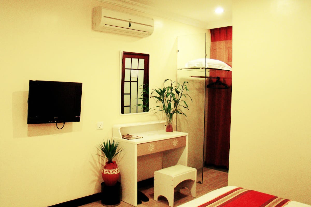 A small, well-lit room with a white desk, a television mounted on the wall, a potted plant, and a red-striped rug.