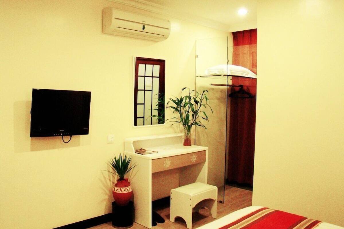 A room with a flat-screen TV, a desk with a chair, a small stool, a potted plant, and a mirror.